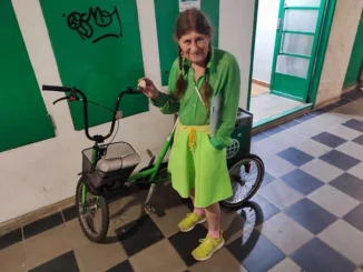 Žena v zeleném oblečení stojí u zelené tříkolky při rozvozu časopisu Metro v pražském domě.