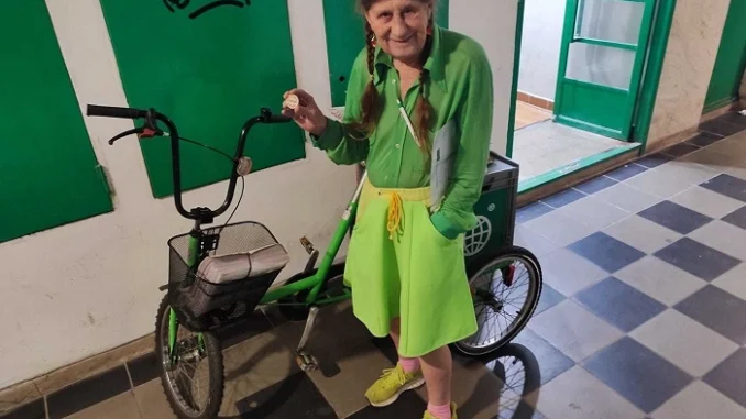 Žena v zeleném oblečení stojí u zelené tříkolky při rozvozu časopisu Metro v pražském domě.