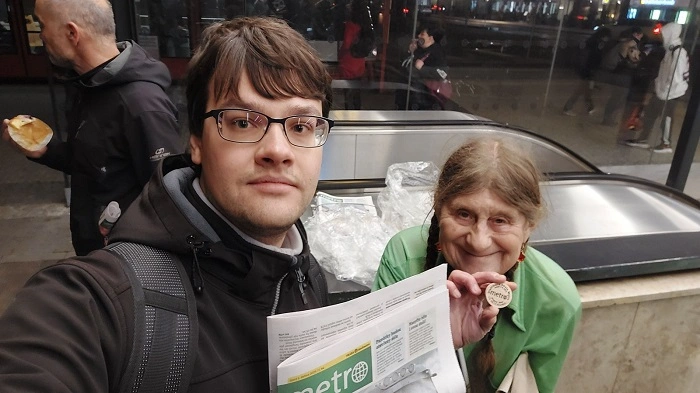 Účastník geocachingového eventu s novinami Metro a ownerkou akce, která drží dřevěný suvenýr z eventu.