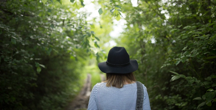 Osoba s kloboukem kráčí lesní cestou obklopenou zelení, symbolizuje objevování a svobodu při hledání keší v geocachingu.
