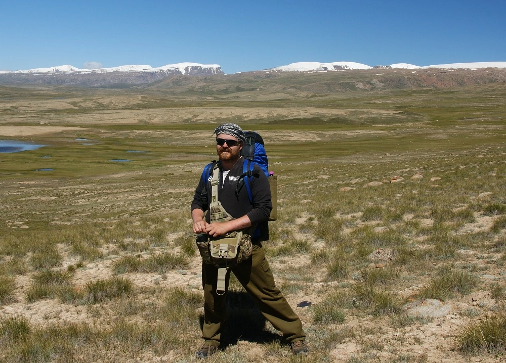 Karamel1 s batohem stojící na travnaté planině v Kyrgyzstánu s výhledem na zasněžené hřebeny.