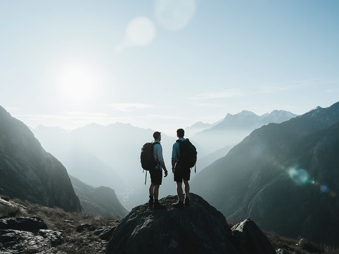 Dva turisté s batohy stojí na skalnatém vrcholu a dívají se na horskou krajinu zalitou ranním sluncem.