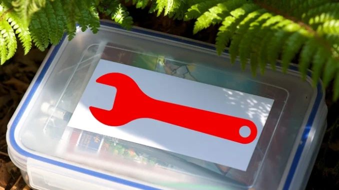 Plastová geocachingová krabička s kartičkou červeného klíče ukrytá pod kapradím – symbol upozornění Owner attention requested.