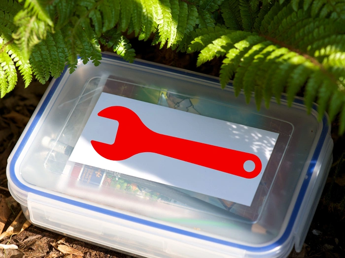 Plastová geocachingová krabička s kartičkou červeného klíče ukrytá pod kapradím – symbol upozornění Owner attention requested.