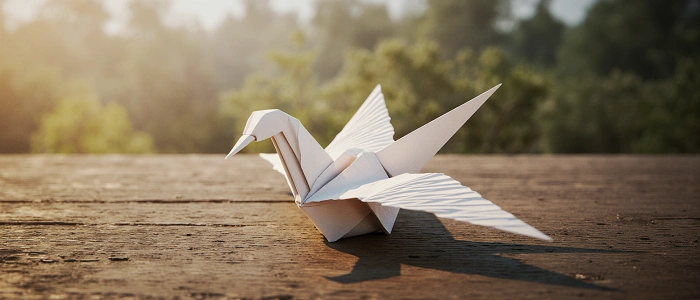 Papírový origami pták složený z bílého papíru leží na dřevěném stole v teplém světle, představuje kolekci Origami zvířátka v geocachingu.