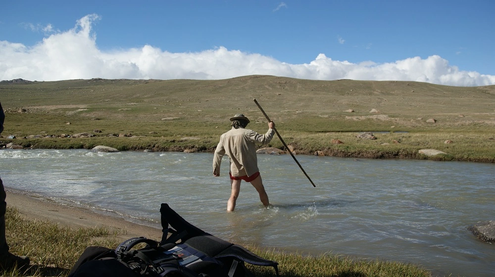 Člověk brodící horskou řeku v otevřené kyrgyzské krajině s pomocí dlouhé hole.