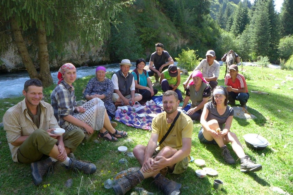Skupina cestovatelů a místních obyvatel sedících u pikniku u horské řeky v Kyrgyzstánu.