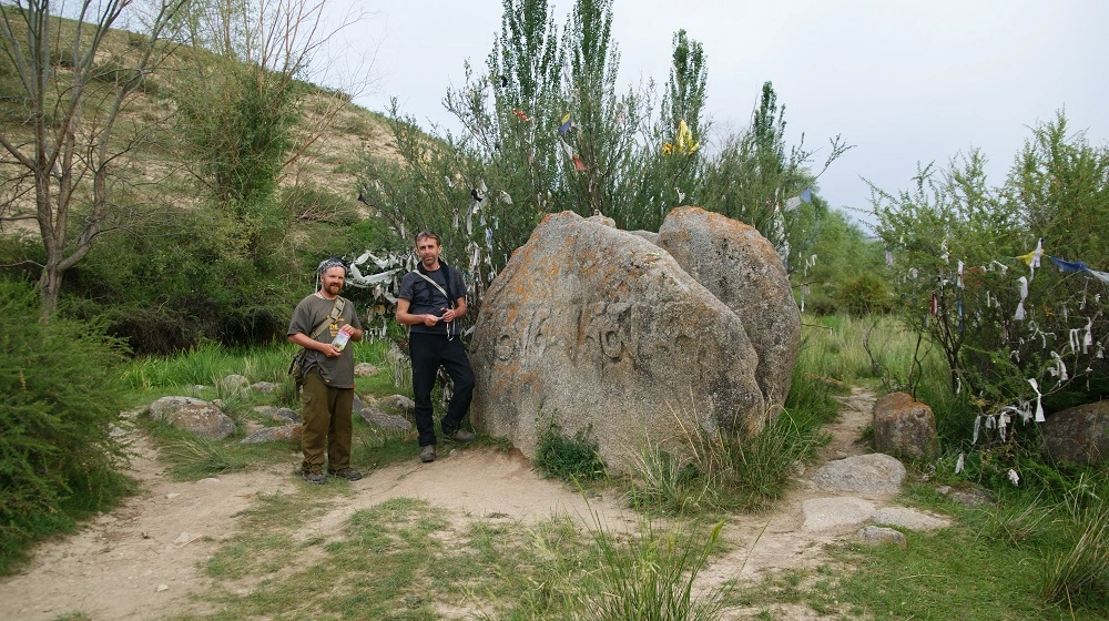 Dva cestovatelé u velkého kamene s rytinami poblíž Issyk-Kul, obklopeni stromy a stezkami.